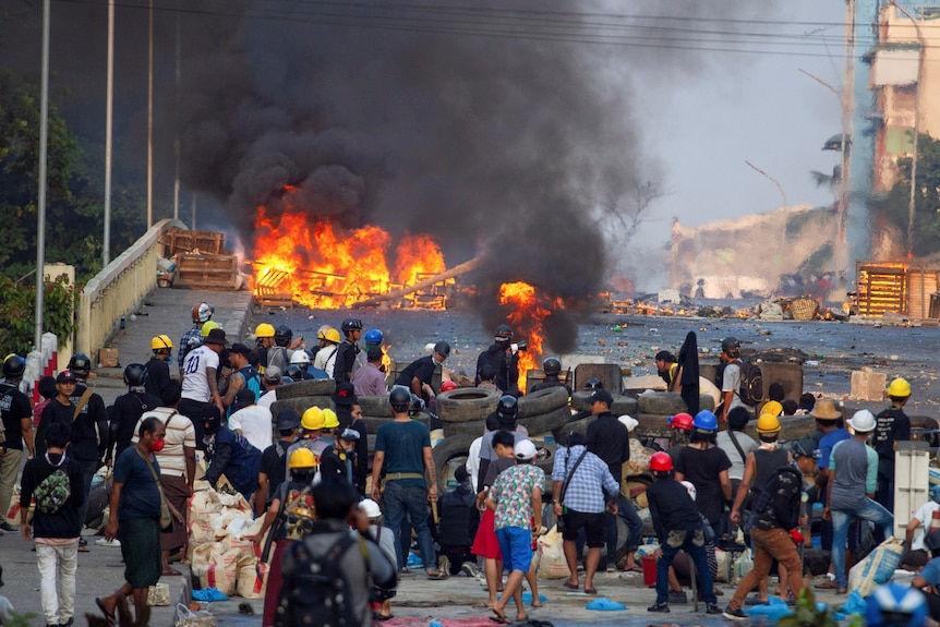 A fire burns in the background on a rundown street with a crowd of protesters in foreground.