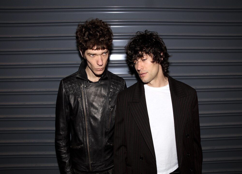 Two men in black jackets and white shirts stand next to each other against a gunmetal grey roller door