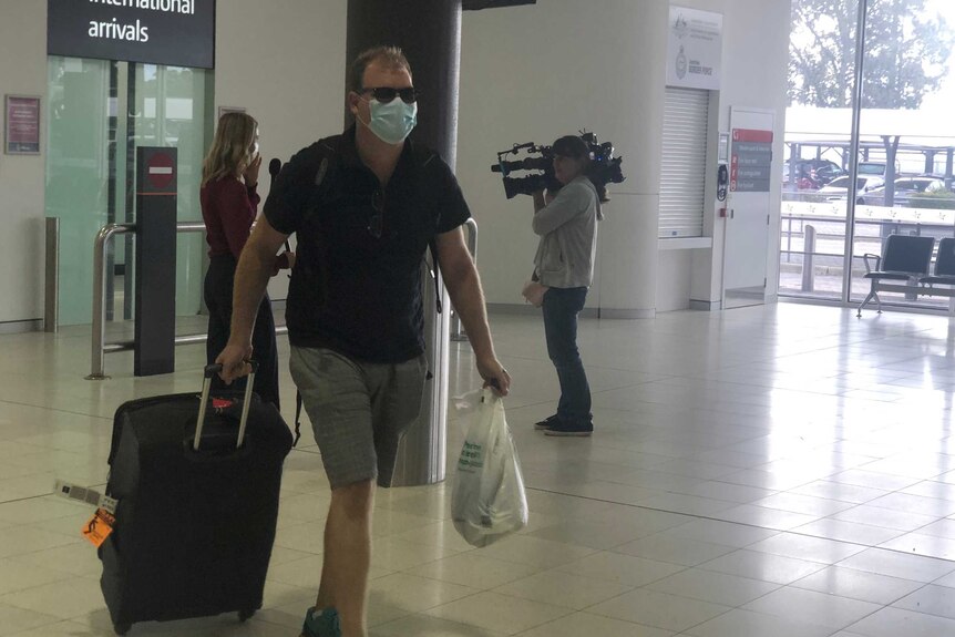 A man walks through an airport holding a suitcase and a bag and wearing a face mask.