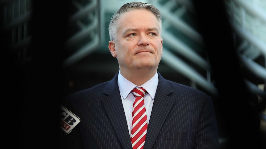 Cormann is seen through a dark blurry foreground standing behind microphones, looking towards right of frame.