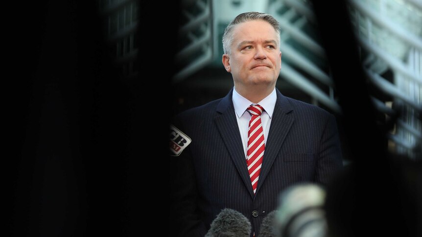 Cormann is seen through a dark blurry foreground standing behind microphones, looking towards right of frame.