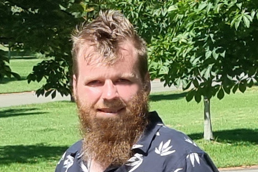 A man with a long blond beard is standing in a green park with trees behind him. He is smiling.