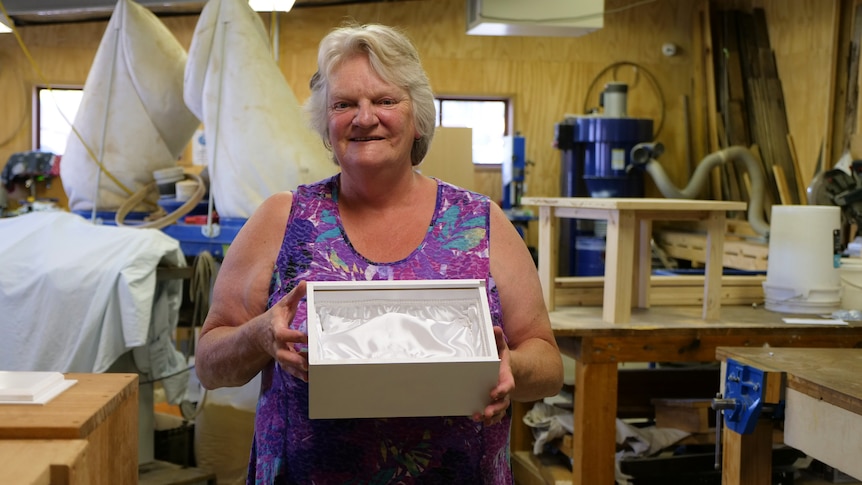 Sue is standing in a woodwork studio. She is holding a white box open to show the satin inside.