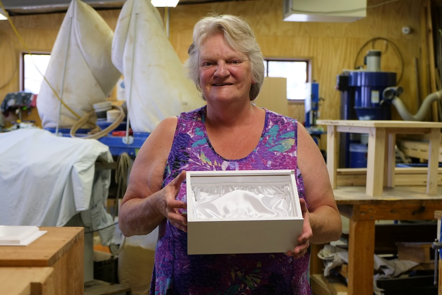 Sue is standing in a woodwork studio. She is holding a white box open to show the satin inside.