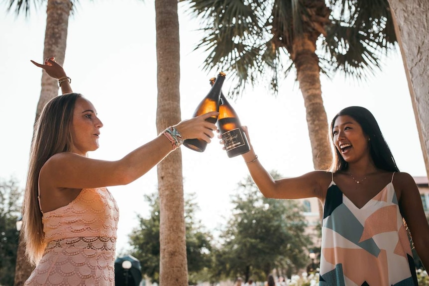 Two women cheers with wine bottles underneath palm trees.
