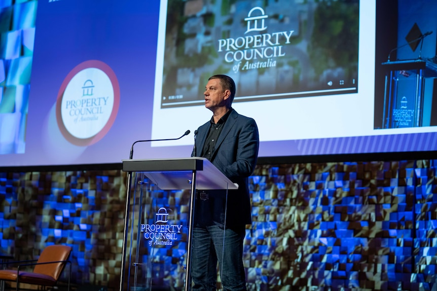 A man addresses an event in a ballroom at night standing at a glass podium 