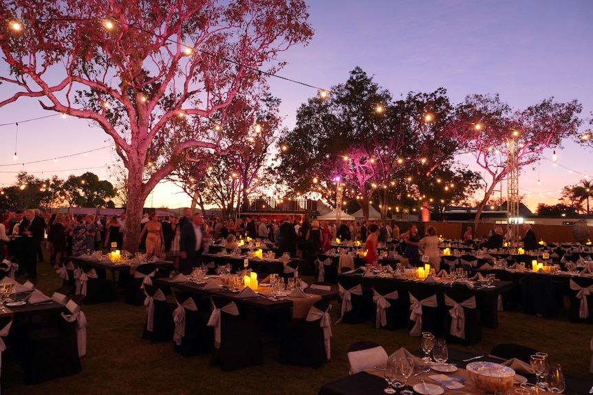 Tables and chairs for a gala dinner are set up outside at night with pink lighting. 