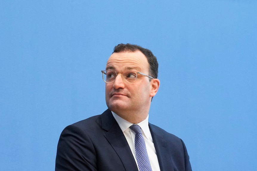 Un hombre con el pelo negro corto en un traje de negocios mira hacia arriba sentado frente a una pared azul 