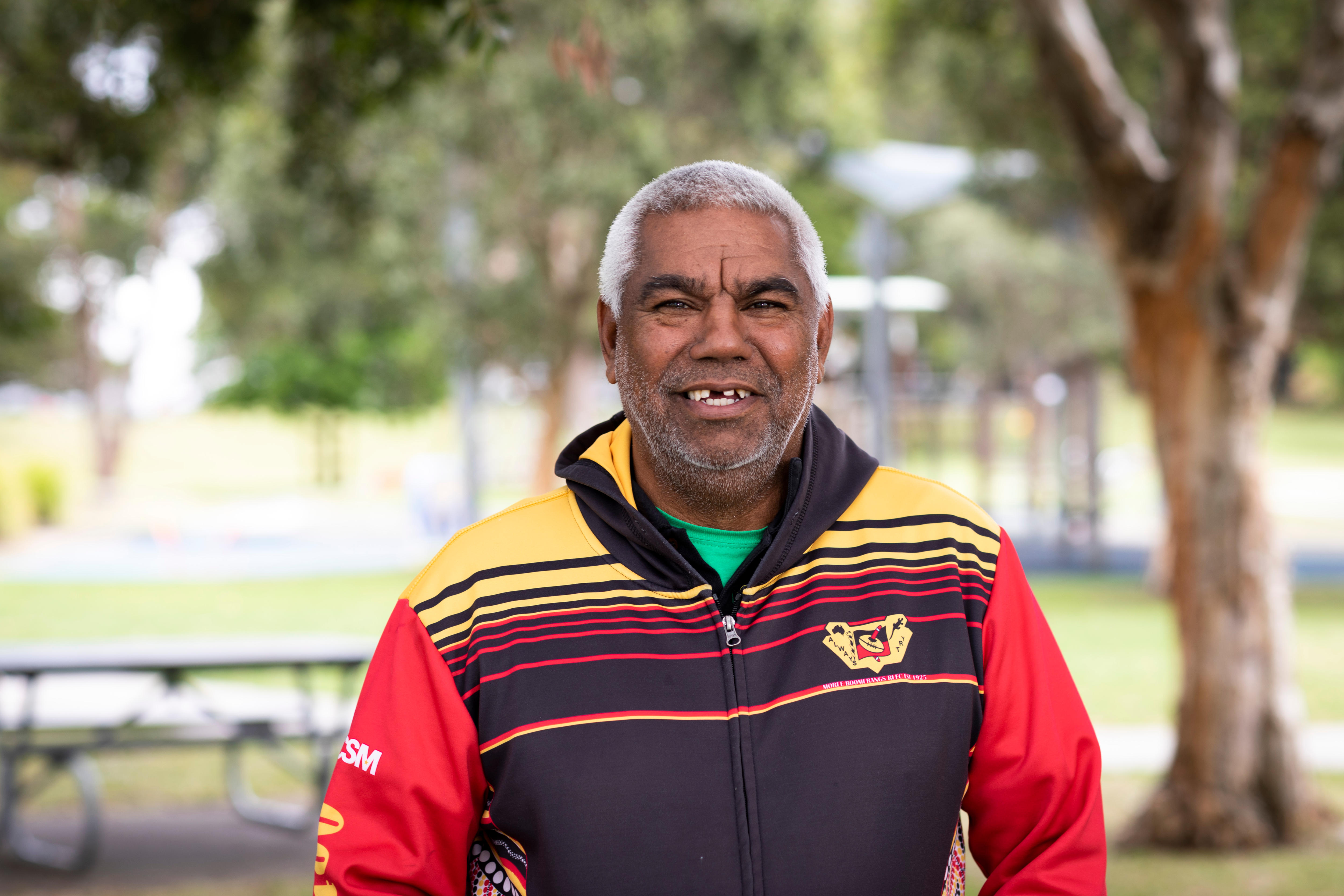 Craig Hammond wears a bright red, black and yellow top and smiles, outdoors.