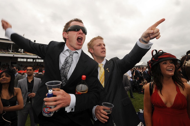 Two men in suits point and yell in front of a crowd of racegoers.