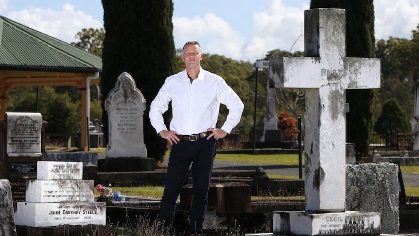 Man in white shirt and black pants stands with his hands on his hips in the middle of a graveyard.