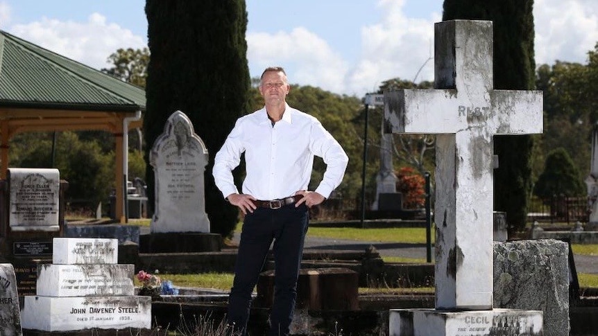 Man in white shirt and black pants stands with his hands on his hips in the middle of a graveyard.