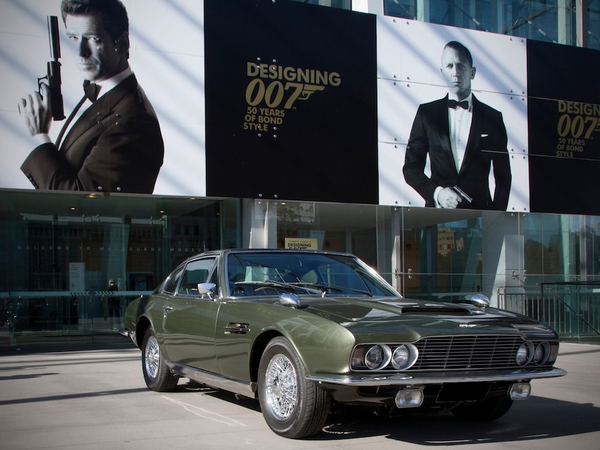 An olive green sports car parked outside a museum with a sign advertising a bond exhibition