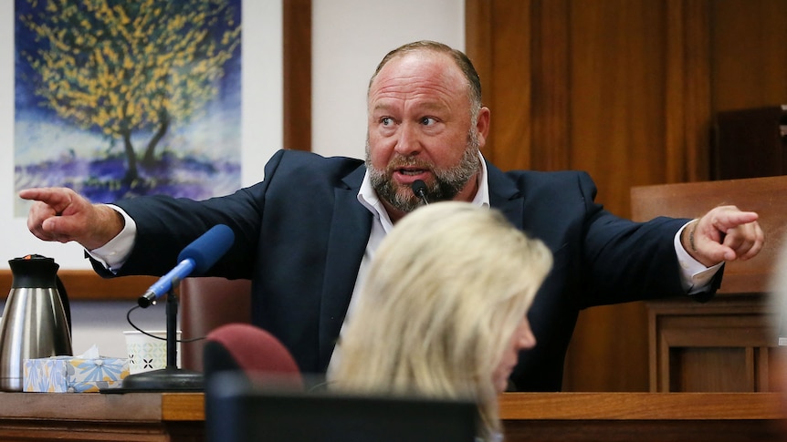 American Alex Jones sits in a courtroom witness stand, wearing a suit and pointing with both hands into the courtroom