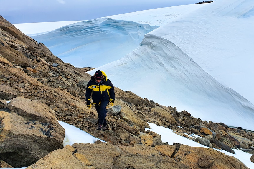 A woman wearing a black and yellow puffer walks up a rocky hill, with glacier behind her.