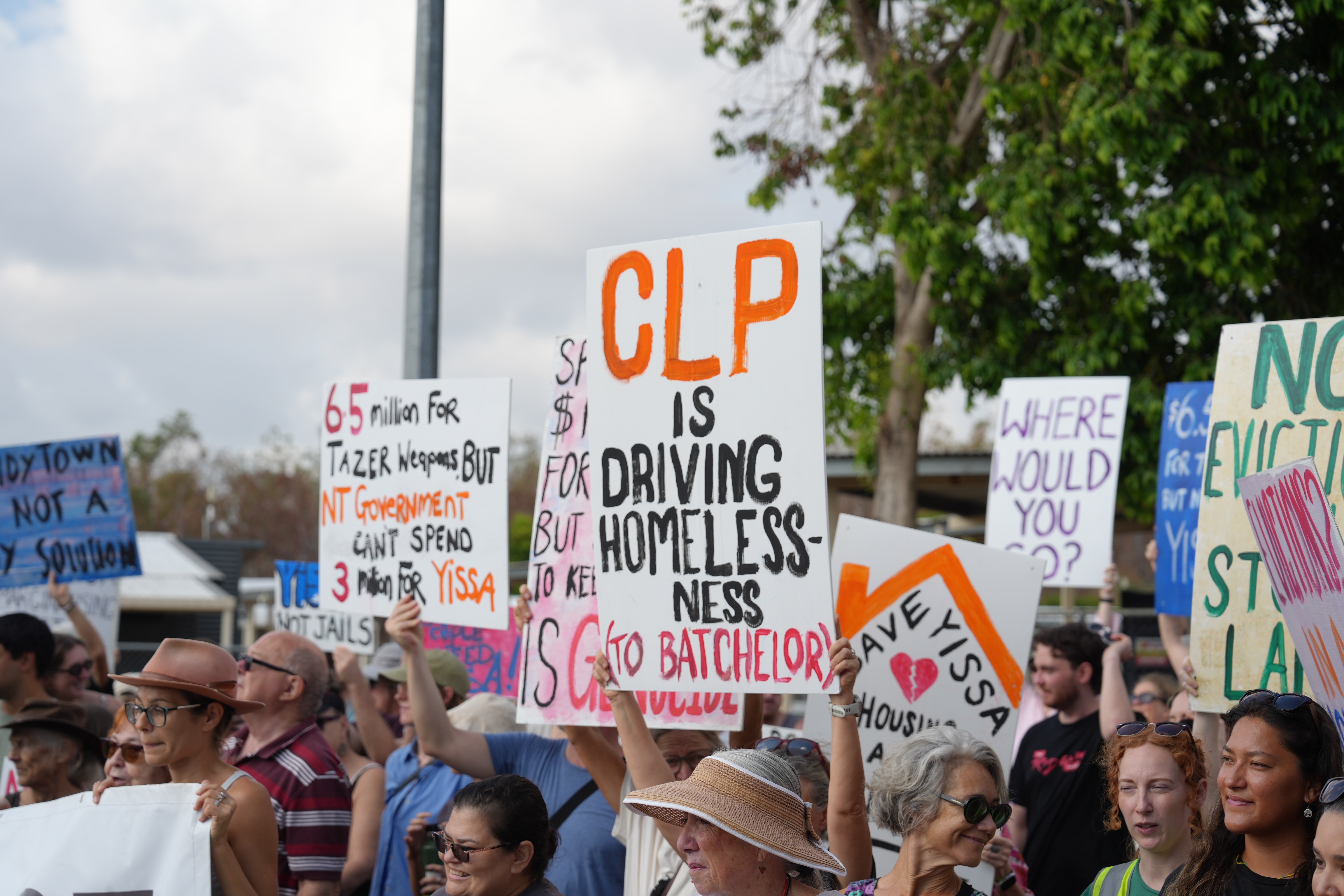A person holds a sign reading "CLP is driving homelessness (to Batchelor)" while surrounded by other protestors.
