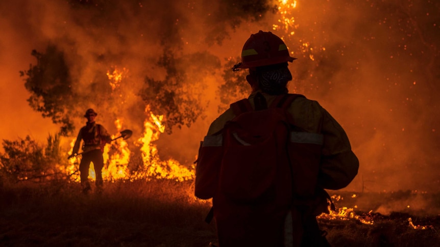 Two firefighters with shovels stand near a fire at night time.