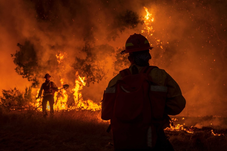 Two firefighters with shovels stand near a fire at night time.