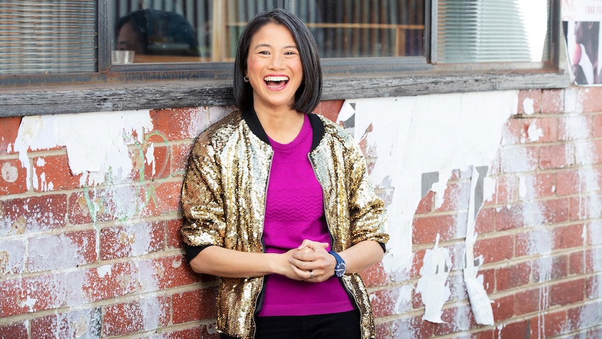 Lisa Leong leaning against a brick wall with windows in it, laughing.