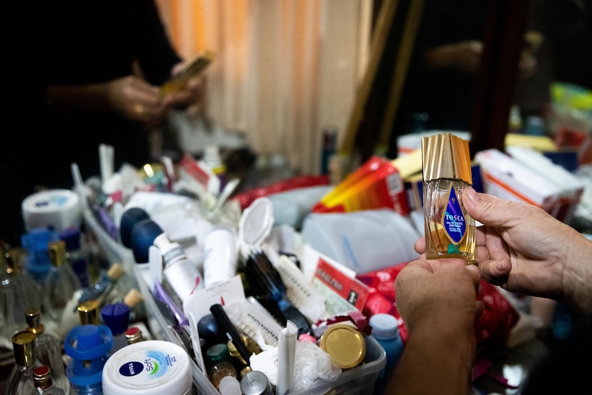 A woman holds a bottle of perfume in front of a cluttered vanity desk.