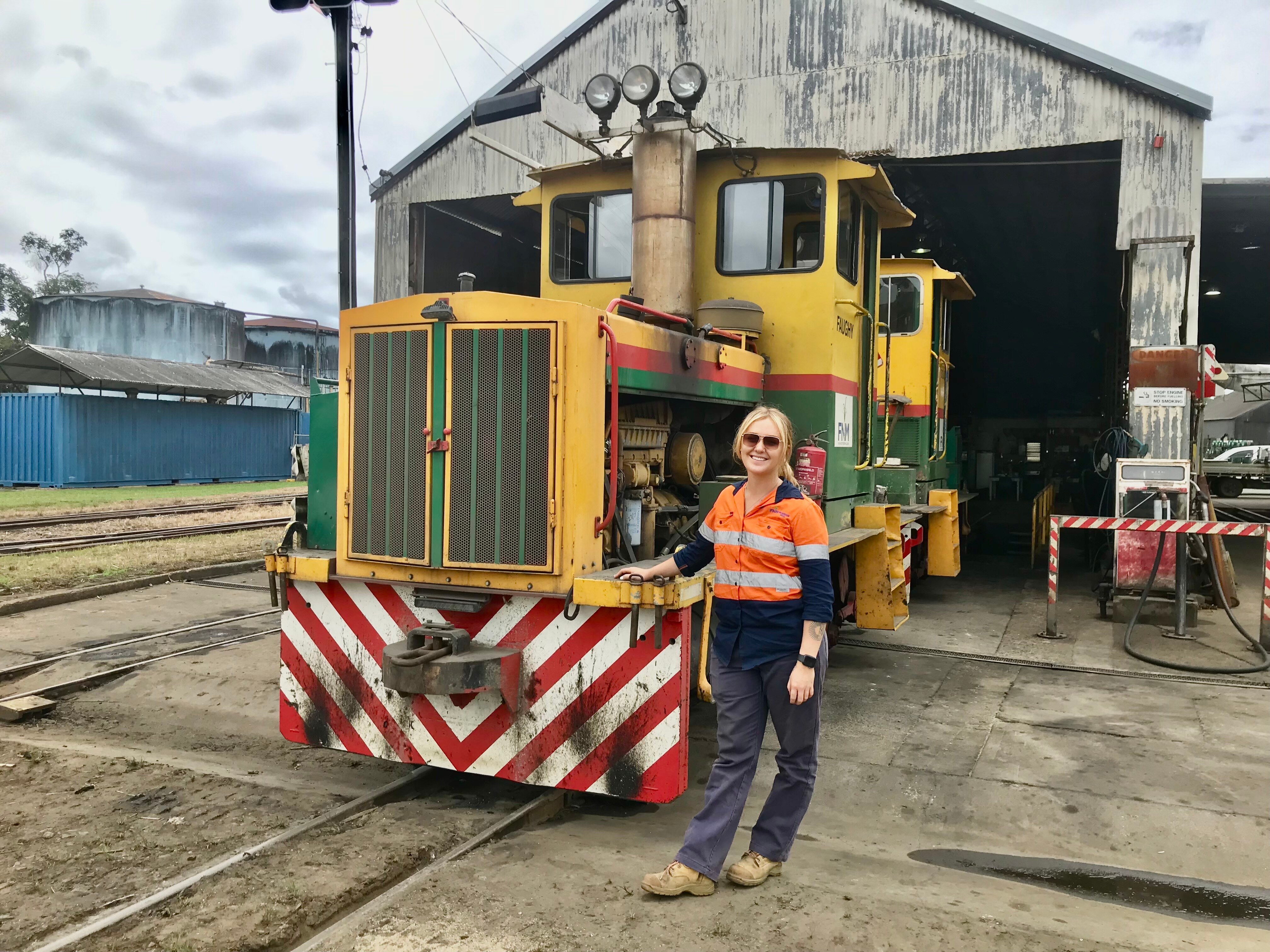 A young woman with a blonde ponytail, high-vis work top and boots, leans on a cane train engine.