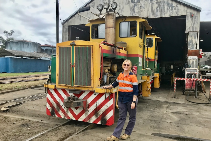 A young woman with a blonde ponytail, high-vis work top and boots, leans on a cane train engine.