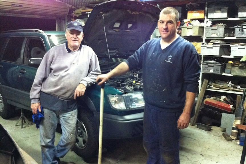 Two men stand in a mechanics garage next to a car with its bonnet up. They smile, oil on their shirts and tools in their hands.