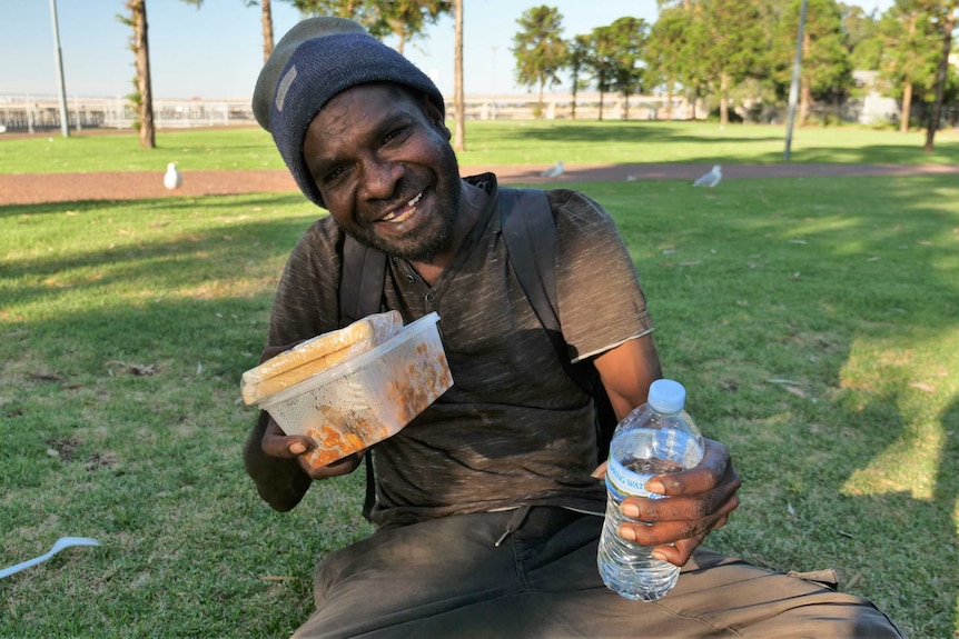 Man smiles at camera holding meal in a plastic box.