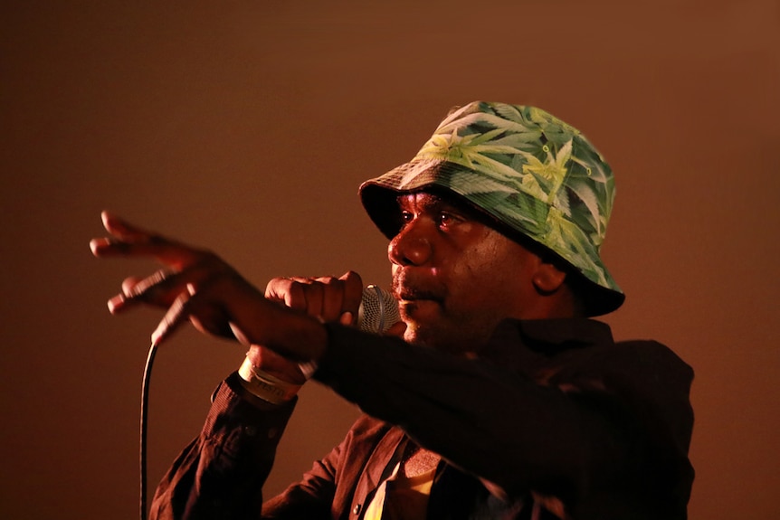 Aborignal man performing on stage, pointing and wearing a green hat