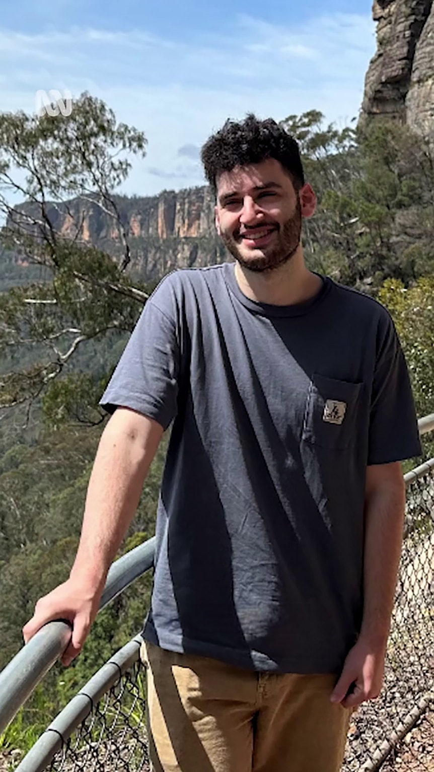 A young man with a beard poses in front of a lookout overlooking bushland.