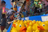 yellow rubber docks bobbing in water, children one side and carnival operator with stuffed toys on other