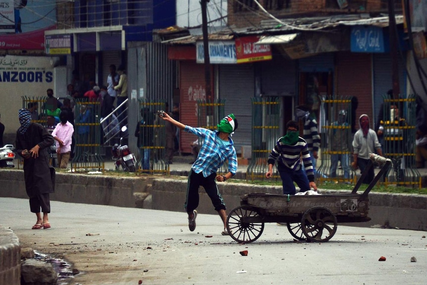 View down a street of a masked protester throwing a large stone.