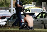 A shirtless man kneels on the ground talking on the phone as a police officer with a rifle walks past.