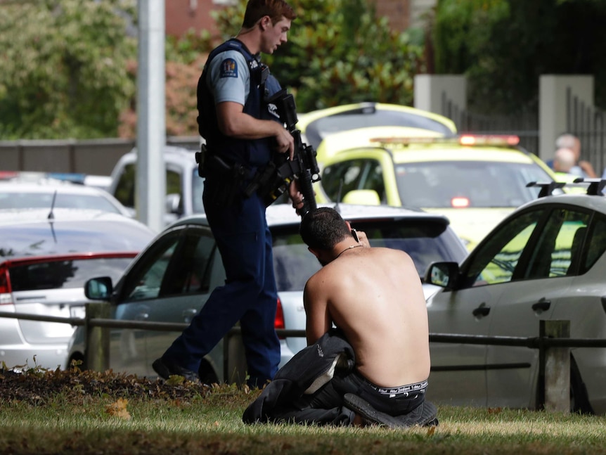 A shirtless man kneels on the ground talking on the phone as a police officer with a rifle walks past.