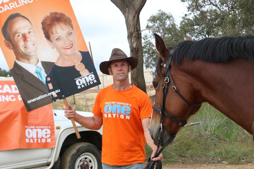A man in an orange One Nation t-shirt stands in front of a One Nation sign with a horse, holding a placard.