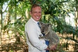 A smiling man in a business shirt cuddles a koala.