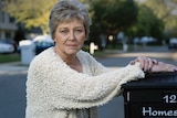 A woman in a cream cardigan with grey cropped hair leans on her mail box