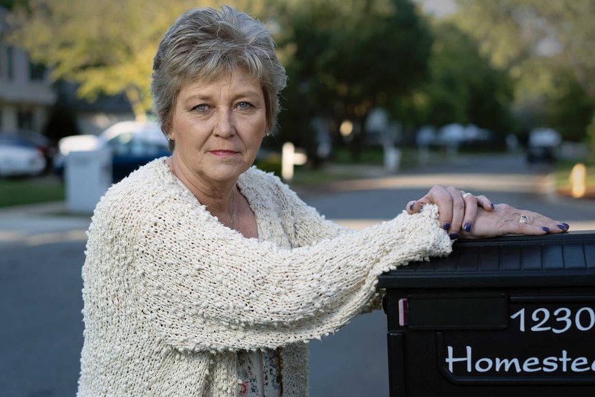 A woman in a cream cardigan with grey cropped hair leans on her mail box