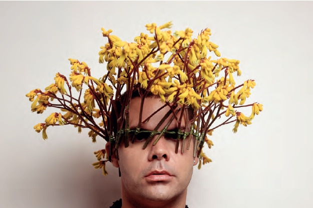 An artistic portrait showing the artist with his face partially obscured by an arrangement of yellow flowers with brown stems.