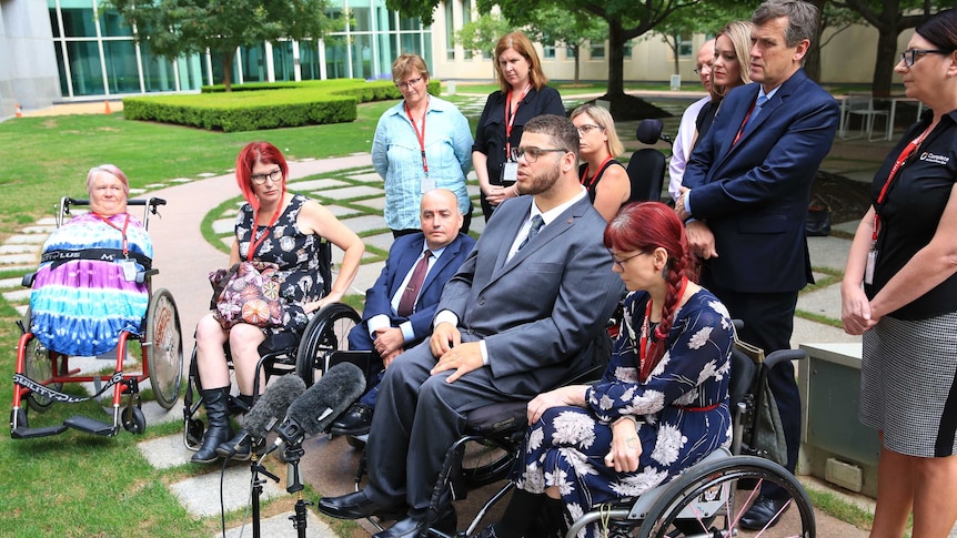 A group of men and women in wheelchairs sit around a microphone at a press conference within a court yard