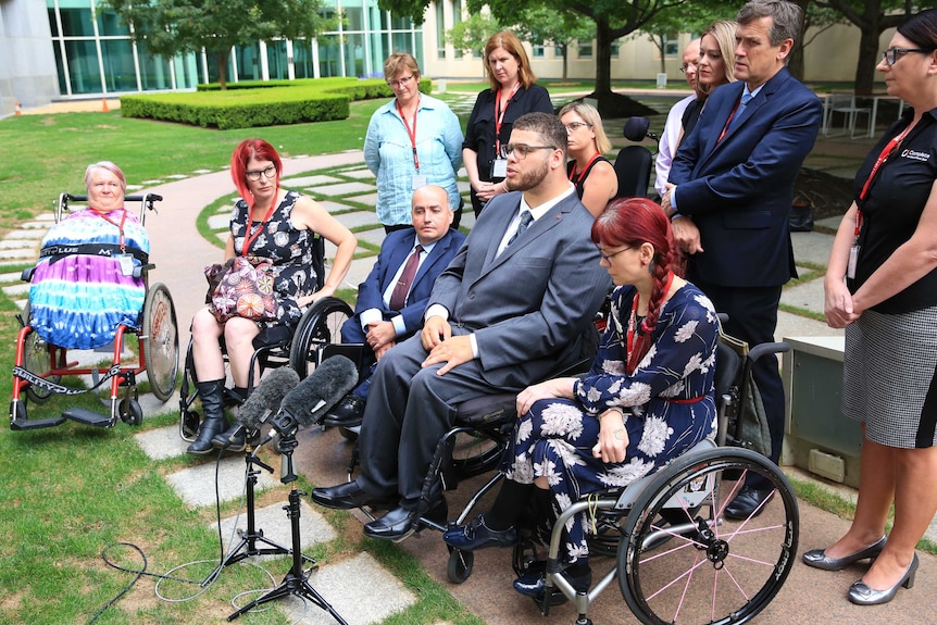 A group of men and women in wheelchairs sit around a microphone at a press conference within a court yard