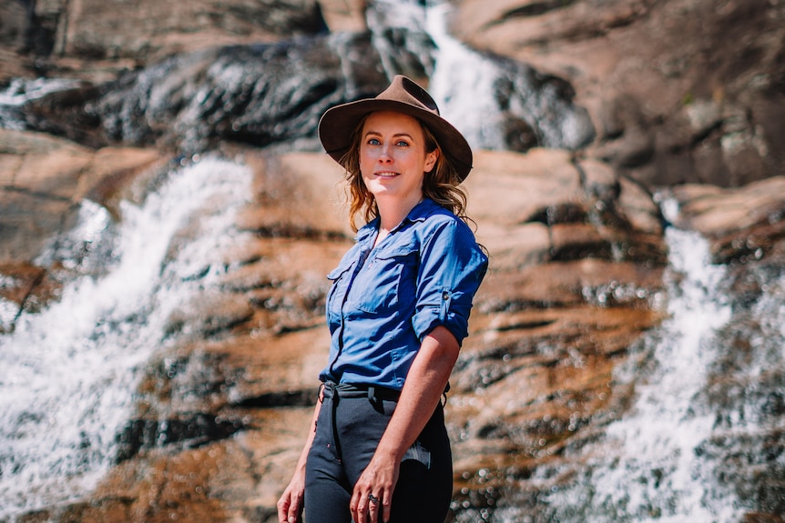 A woman wearing a blue shirt and a brown hat stands in front of a rockface