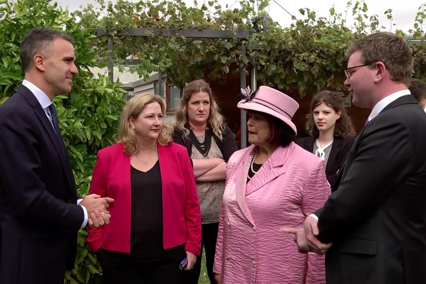 Two men wearing dark suits speak to a woman wearing a red jacket and a woman wearing a pink outfit and hat