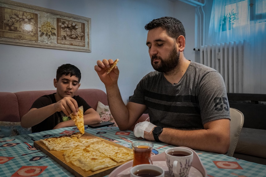 A man and boy sit at a table sharing a plate of flatbread. A tray with cups of tea is also on the table