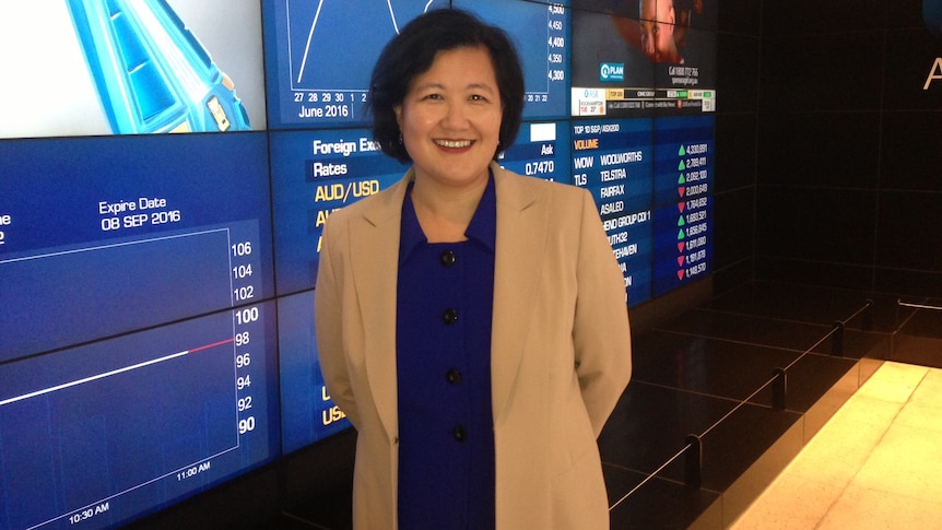 Ming Long standing in front of plasma screens displaying stocks.