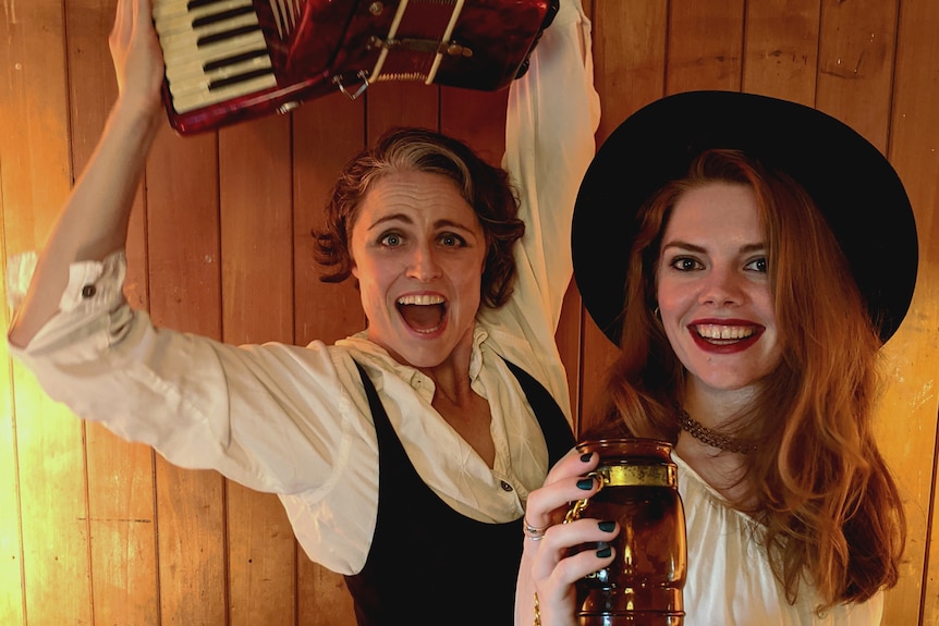 Two women smiling in pirate-themed clothing, one holding an accordion over her head and the other holding a jar.