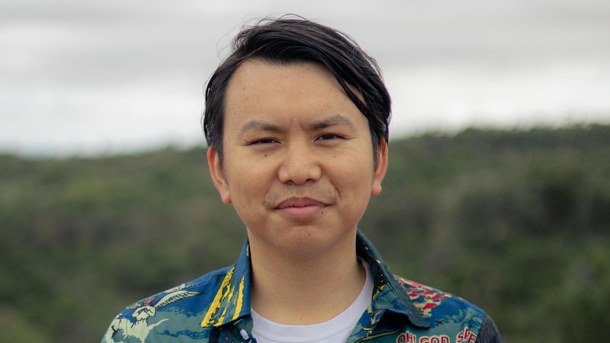 A young man stands outdoors wearing a patterned shirt. 