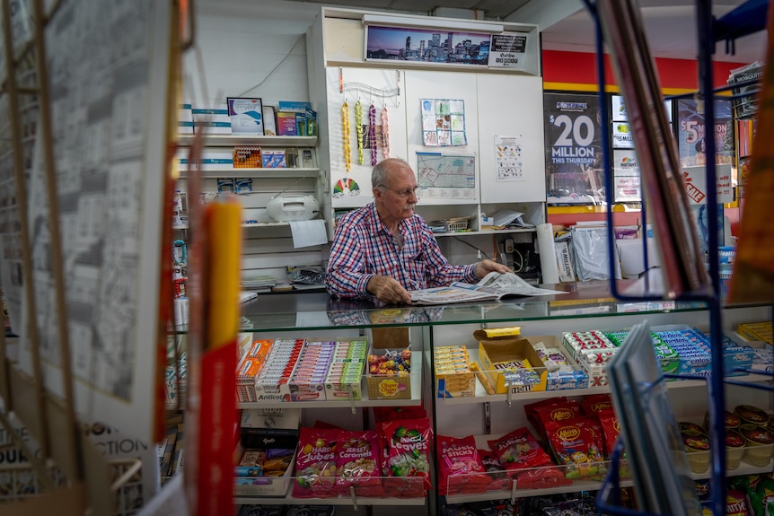 A man sits behind a newsagent's counter and reads a newspaper.