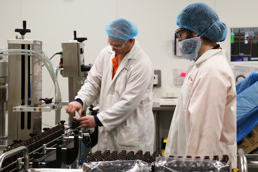 Workers in white coats and hair nets put caps on small brown bottles.
