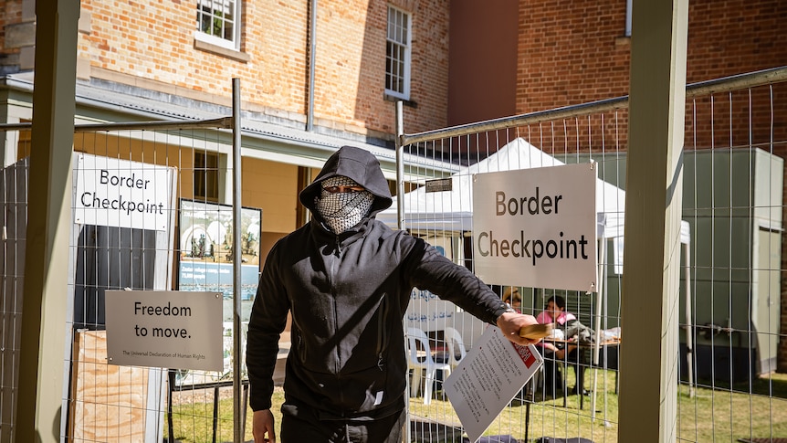 A man wearing a black hoodie and a bandana hiding most of his face stands by a gate with a sign that says Border Checkpoint